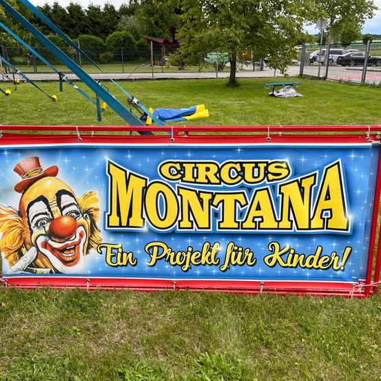 Ein Banner für den Circus Montana ist auf einem Rasenfeld zu sehen. Es zeigt einen Clown mit roter Nase, umgeben von buntem Text.