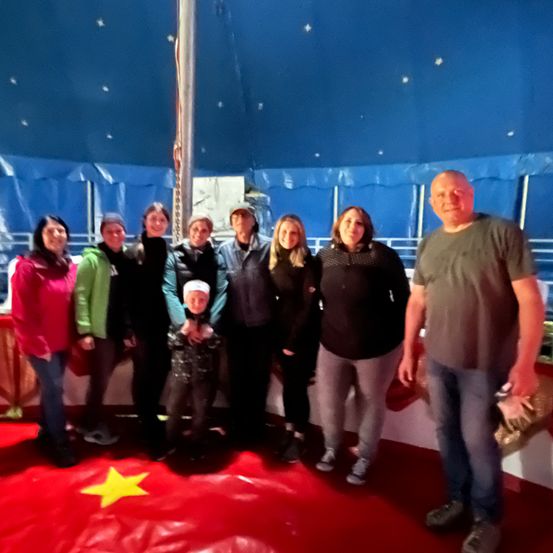 Eine Gruppe von Erwachsenen und einem Kind steht in einem Zelt mit blauer Decke und rotem Boden und posiert für ein Foto. Sie sind in Winterkleidung gekleidet.