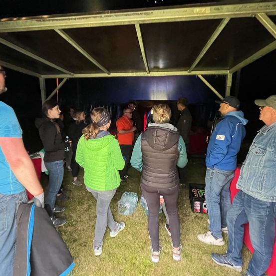 Eine Gruppe von Menschen, darunter Frauen in Jacken und Männer in Mützen, stehen und unterhalten sich unter einem Zelt bei Nacht. Einige halten Flaschen.