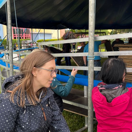 Eine Gruppe von Menschen baut eine Struktur unter einem Zelt auf. Eine Frau mit Brille und einer gepunkteten Jacke lächelt.