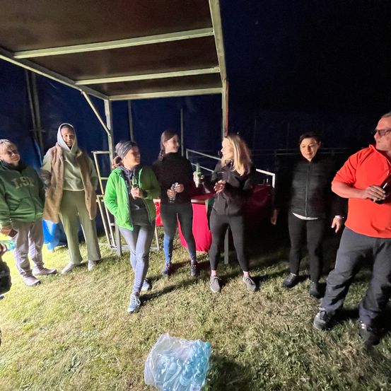 In der Nacht stehen eine Gruppe von Menschen in einem Außenbereich unter einem Zelt. Einige halten Getränke, andere stehen vor einem Tisch. Der Bereich ist von Gras umgeben.