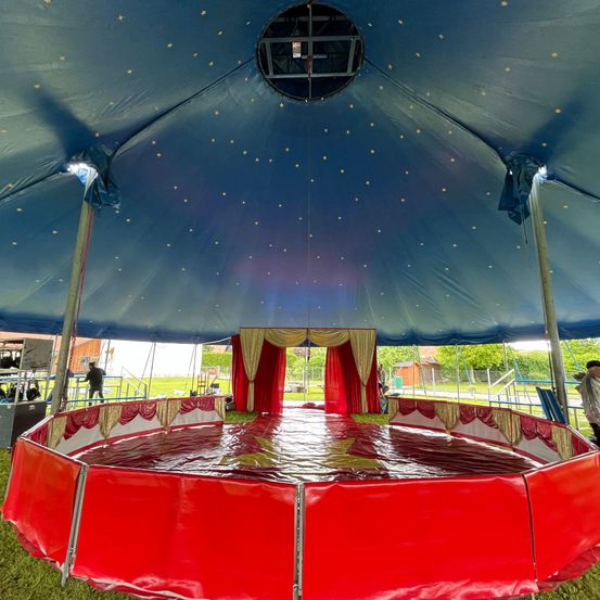 Ein Zirkuszelt ist aufgebaut mit einem roten Ring auf dem Boden, umgeben von roten Barrieren. Das Zelt ist blau mit gelben Sternen und einer kreisförmigen Öffnung. Leute bereiten den Bereich vor.