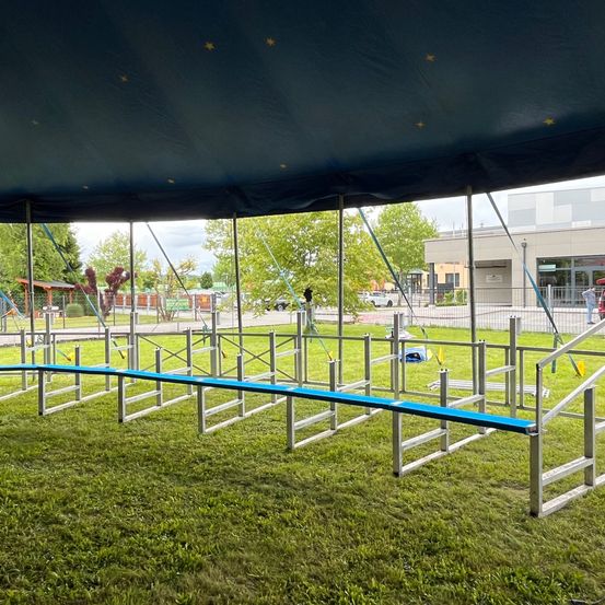 Ein Außenbereich mit einem großen Zelt und einer Reihe von Metallhindernissen auf einem Rasenfeld, mit Bäumen und einem Gebäude im Hintergrund.