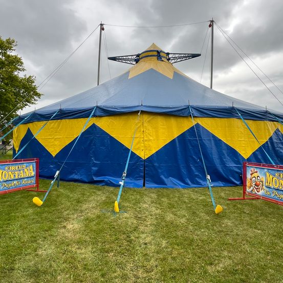 Ein großes blau-gelbes Zirkuszelt steht auf einem Rasenfeld, mit zwei Bannern auf beiden Seiten. Ein Banner sagt 'Circus Montana' und das andere zeigt einen Clown.