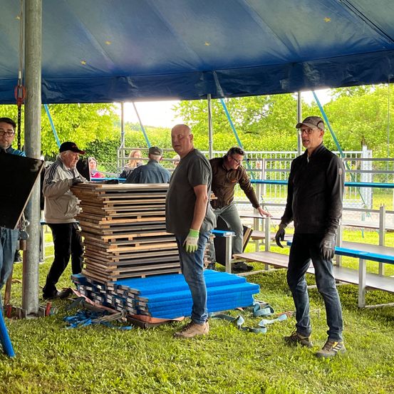 Eine Gruppe von Männern arbeitet unter einem blauen Zelt. Sie stehen auf einem Rasenfeld mit mehreren Holzpaletten und blauen Matten. Ein Mann lehnt sich an ein Metallgitter.