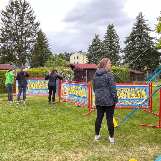 Vier Personen stehen in der Nähe einer Circus Montana-Aufstellung auf einem Rasenfeld. Sie sind in der Nähe eines bunten Schildes und eines Zauns. Eine Frau schaut in die Kamera. Das Gebiet ist von Bäumen und einem Haus umgeben.