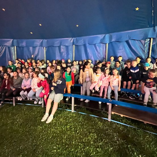 Eine große Gruppe von Kindern ist in einem Zelt versammelt, um eine Aufführung zu sehen, einige sitzen auf blauen Bänken. Das Zelt ist blau mit gelben Sternen.