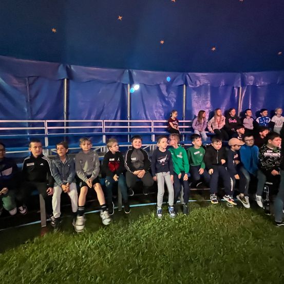 Eine Gruppe von Kindern und Jugendlichen sitzt unter einem blauen Zelt mit Sternen. Sie sitzen auf Bänken, einige tragen Jacken und Turnschuhe. Der Boden ist grasbedeckt.