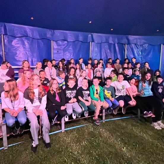 Eine Gruppe von Kindern und Teenagern sitzt in einem Zelt unter einer blauen Plane. Sie lächeln und scheinen eine Veranstaltung zu genießen.