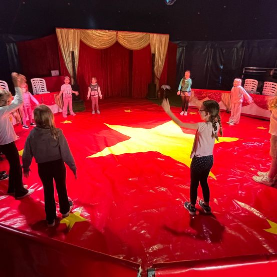 Eine Gruppe von Kindern tritt auf einer roten Zirkusbühne mit einem großen gelben Stern auf. Sie tragen Freizeitkleidung und einige haben Brillen. Hinter ihnen sind rote Vorhänge und ein Tisch zu sehen.