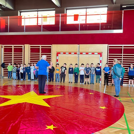 Ein Mann steht in einer Turnhalle mit einem roten Kreis mit einem gelben Stern. Kinder stehen hinter ihm und schauen zu. Eine Frau steht rechts und schaut auf den Mann.