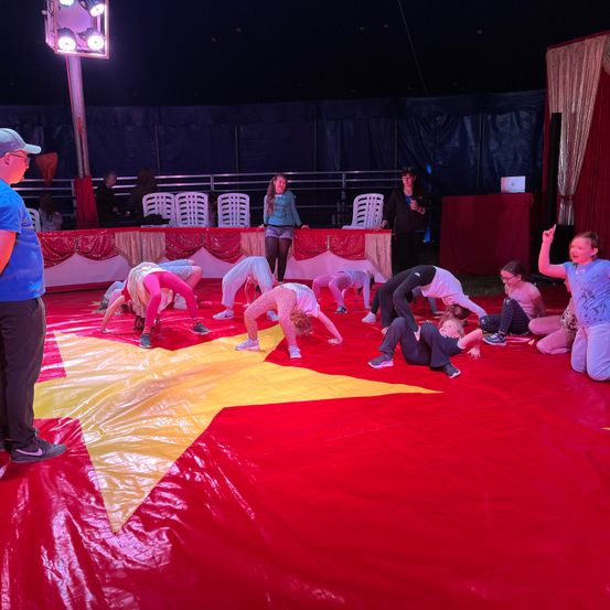 Kinder führen Akrobatik auf einem roten Teppich unter einem Zelt vor. Eine Frau steht hinter ihnen, und das Publikum schaut zu. Das Zelt ist mit rotem Stoff und Vorhängen geschmückt.