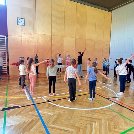 Eine Gruppe von Kindern führt eine Übungsroutine in einer Turnhalle durch. Eine Frau führt sie an, während sie in der Mitte steht. Der Boden ist mit Linien markiert und die Wände sind golden.