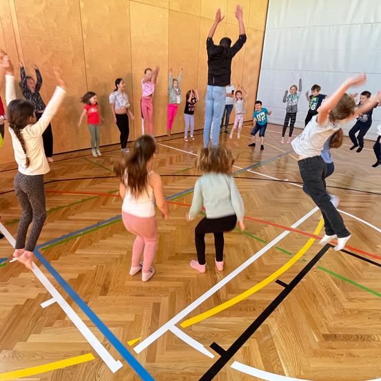 In einer Turnhalle führen Kinder eine koordinierte Übungsroutine durch. Ein Mann steht in der Mitte, und die Kinder bilden einen Kreis um ihn und heben ihre Arme.