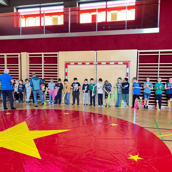 In einer Turnhalle stehen eine Gruppe von Kindern in einem Kreis um eine rote Matte mit einem gelben Stern. Zwei Erwachsene stehen in der Nähe, einer hält ein buntes Band. Die Kinder halten Bänder, und die Turnhalle hat eine rote Wand und eine Glaswand.