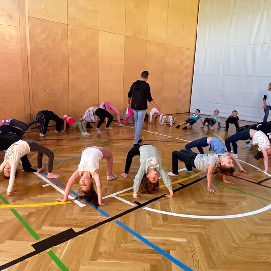 Eine Gruppe von Kindern in verschiedenen Posen bildet einen Kreis auf einem Holzboden, angeführt von einem Mann in der Mitte. Der Raum hat Holzwände und einen weißen Vorhang im Hintergrund.
