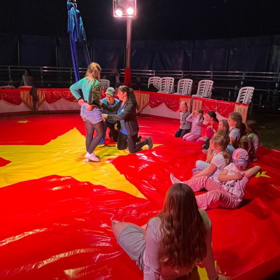 Eine Gruppe von Kindern und Erwachsenen versammelt sich um einen Zirkusring. Eine Frau wird von einem Erwachsenen unterstützt, während sie auf einem roten und gelben Teppich steht. Die Kinder sitzen und schauen zu.