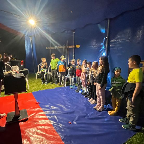 In einem Zelt stehen Kinder auf einem blauen Teppich unter einer Lampe und lauschen einem Erwachsenen auf der Bühne.