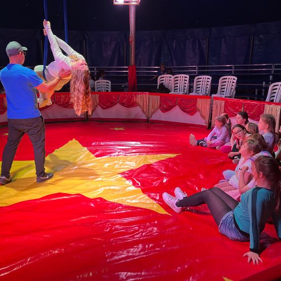Ein Mann in einem blauen T-Shirt hilft einer Frau in weißen Hosenträgerhosen bei einem Zirkusakt. Zuschauer, darunter Kinder, schauen von roten gepolsterten Sitzen zu. Der Ring ist mit einem rot-gelben Planen bedeckt.