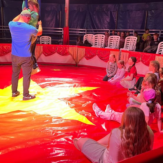 Ein Mann hilft einem Kind, an einem Trapez in einem Zirkus zu hängen. Zuschauer sitzen auf dem Boden und schauen zu. Ein roter und gelber Teppich bedeckt den Boden.