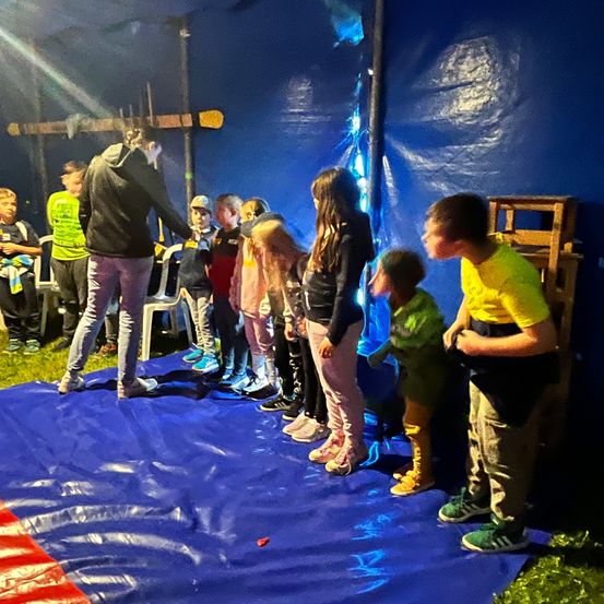 Eine Gruppe von Kindern steht in einer Reihe in einem blauen Zelt, wahrscheinlich für eine Aufführung. Das Zelt hat einen Holzboden und ist mit blauer Plastik bedeckt. Die Kinder tragen Turnschuhe.