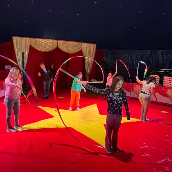 Eine Gruppe von Kindern übt mit bunten Bändern in einem Zirkuszelt, unter einem sternförmigen Scheinwerfer.