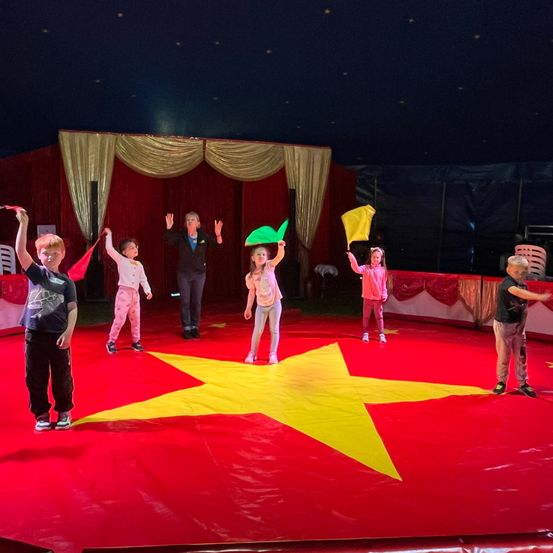 Eine Gruppe von Kindern und einer Frau steht auf einer Zirkusbühne, hält Flaggen und hat einen roten Boden mit einem gelben Stern in der Mitte.