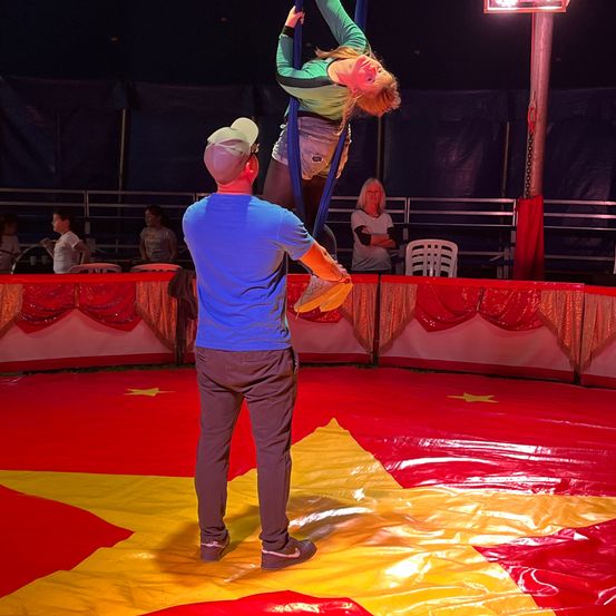 Ein Artist hängt kopfüber von einem Trapez ab, gestützt von einem Mann in einem Zirkuszelt, während Zuschauer auf den Tribünen zuschauen.