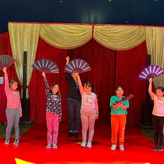 Fünf Kinder stehen auf der Bühne und halten LED-Ventilatoren, während ein Mann hinter ihnen steht. Die Bühne ist mit roten und goldenen Vorhängen bedeckt.