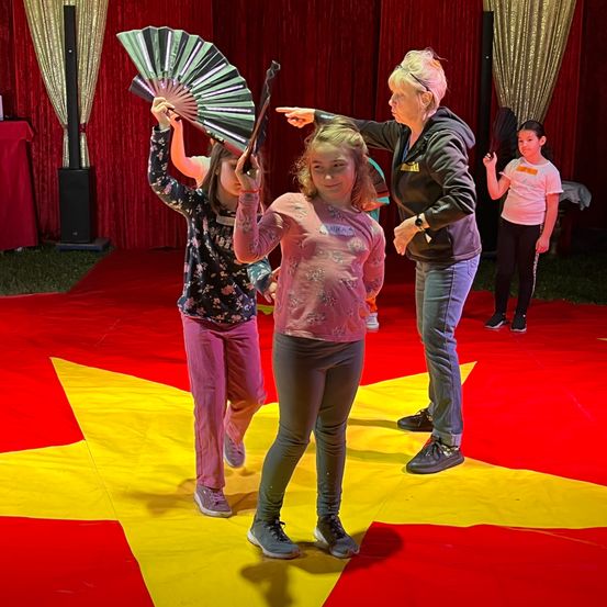 Drei Mädchen auf einem roten Teppich, eines hält einen Fächer, das andere einen Stock. Sie werden von einer erwachsenen Frau geführt. Hinter ihnen ist ein roter Vorhang.