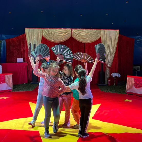 Eine Gruppe von Kindern auf einer Zirkusbühne mit einem rot-gelben Hintergrund, die Fächer hochhalten, mit einer sternübersäten Decke.