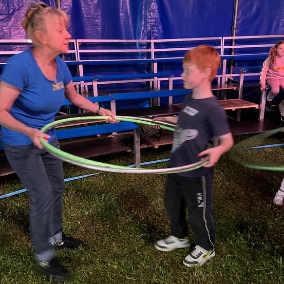 Eine Frau hilft einem jungen Jungen, einen Hula-Hoop unter einem blauen Zelt zu benutzen. Dahinter sitzen leere Bänke und ein Mädchen.