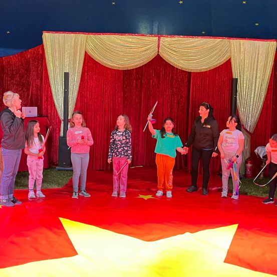 Eine Gruppe von Kindern und Erwachsenen steht auf einem roten Teppich, wahrscheinlich in einem Zirkus. Sie halten Zauberstäbe, und zwei Frauen führen sie an. Hinter ihnen befindet sich ein roter Hintergrund mit goldenen Vorhängen.