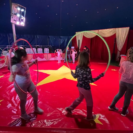 Eine Gruppe von Kindern übt Hula-Hoop in einem Zirkuszelt mit rotem Boden und einem gelben Stern. Ein Erwachsener ist in der Nähe.