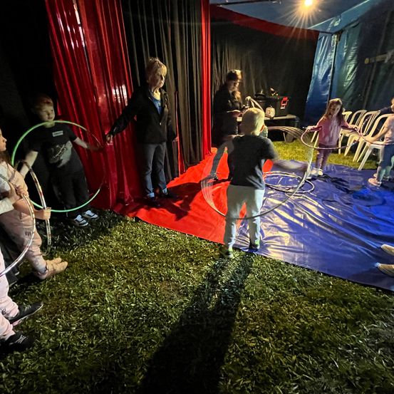 Eine Gruppe von Kindern und Erwachsenen übt Hula-Hoopen in einem Zirkuszelt unter blauem Himmel.