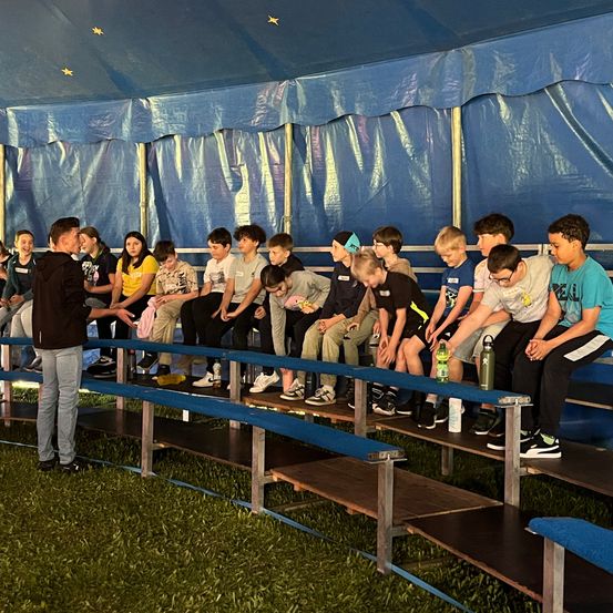 Eine Gruppe von Kindern sitzt auf Tribünen unter einem blauen Zelt und hört einem Mann zu, der vor ihnen steht. Der Mann spricht und die Kinder schauen ihn an.