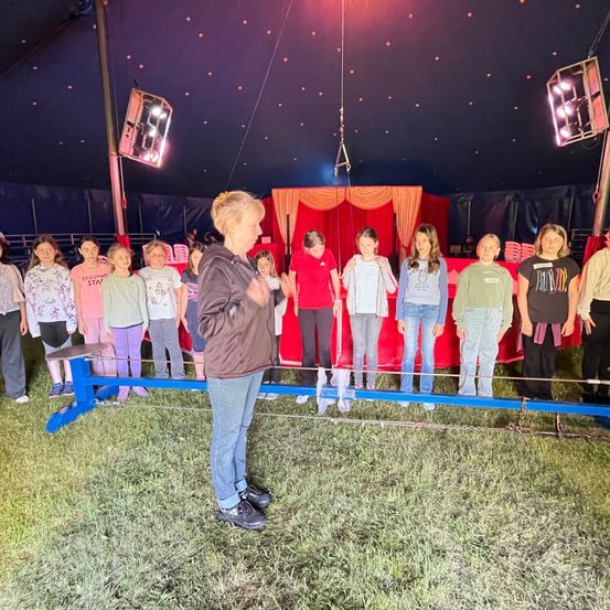 Eine Frau steht vor einer Gruppe von Kindern in einem Zirkuszelt und hält eine Schnur. Die Kinder stehen in einer Reihe auf einer blauen Stange und tragen verschiedene Freizeitkleidung. Scheinwerfer hängen von der Decke.