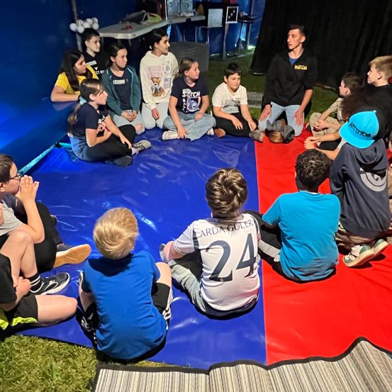 Eine Gruppe von Kindern und einem Mann sitzen auf dem Boden. Einige tragen Turnschuhe. Sie hören dem Mann zu, der steht.