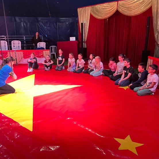 Kinder sitzen auf einem roten Boden in einem Zelt mit einem Stern, Erwachsene beobachten von der Seite, und eine Frau beobachtet sie.