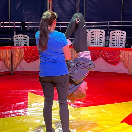 Eine Frau in einem blauen Shirt unterstützt eine andere Frau in schwarzen Leggings bei einer Trapez-Vorführung, die auf einem roten und gelben Mattenboden kopfüber hängt.