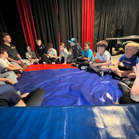 Eine Gruppe von Kindern und einem Mann sitzen im Kreis auf einer blauen Matte. Sie nehmen wahrscheinlich an einem Workshop teil. Dahinter befindet sich eine rote Matte und schwarze Vorhänge.
