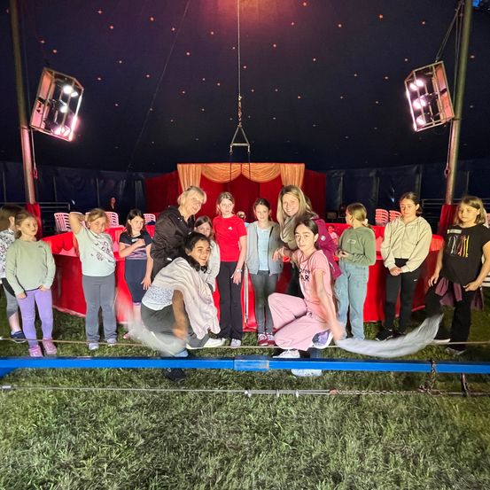 Eine Gruppe von Kindern und Erwachsenen steht in einem Kreis um eine Seilbahn in einem Zirkuszelt. Eine Frau kniet auf dem Seil, während eine andere Frau einen Schwanz hält. Dahinter ist eine rote Bühne mit Vorhängen.