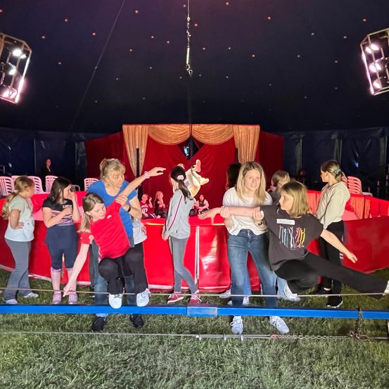 Eine Gruppe junger Mädchen übt das Balancieren auf einem Seil unter einem Zirkuszelt. Ein Mädchen in einem roten Shirt kniet, während sich ein anderes in Schwarz nach vorne streckt. Eine Frau führt sie von hinten an.