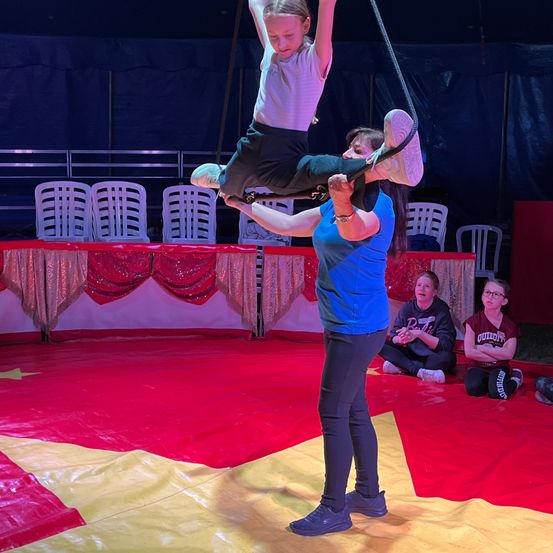 Ein Mädchen führt einen Trapezakt in einem Zirkus mit einer Frau durch, die sie unterstützt. Zuschauer beobachten von den Seitenlinien.
