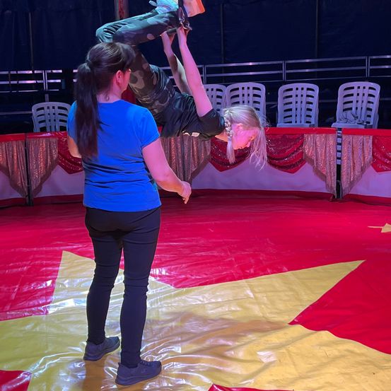 Ein Mädchen führt einen Handstand in einem Zirkuszelt durch. Eine erwachsene Frau hält ihre Beine. Rot und gelbe Matten bedecken den Boden. Stühle stehen im Hintergrund.