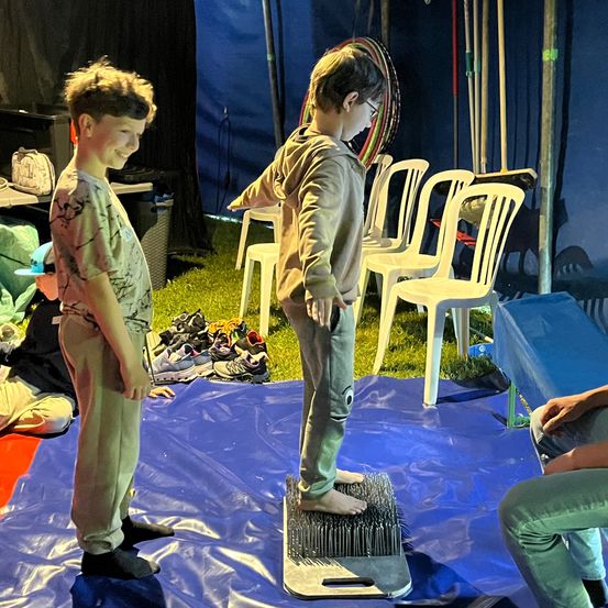 Zwei Jungen üben ihre Balancefähigkeiten auf einem spitzen Brett. Sie stehen barfuß auf dem Brett mit den Händen auf den Hüften. Dahinter stehen Stühle, ein Tisch und eine blaue Plane.