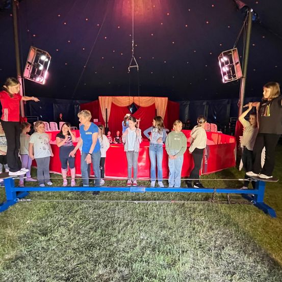 Eine Gruppe von Kindern balanciert auf einem Seil unter einem Zirkuszelt, mit einer roten Bühne im Hintergrund und Scheinwerfern über ihnen.