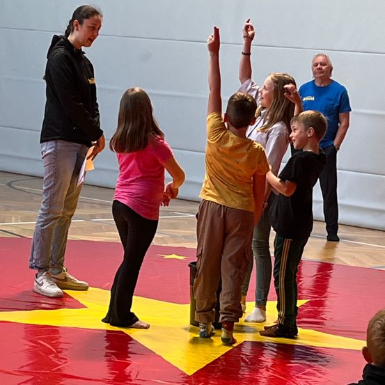 Eine Gruppe von Kindern und einem Mann ist in einer Turnhalle. Die Kinder heben ihre Hände. Der Mann trägt eine schwarze Jacke und Turnschuhe. Die Kinder tragen Freizeitkleidung.