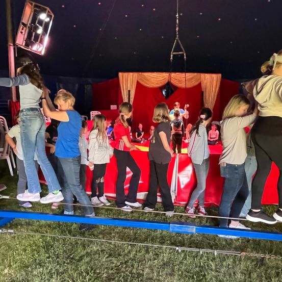 Kinder in einem Zirkuszelt gehen auf einem Seil, während andere von einer rot drapierten Bühne zusehen. Eine Scheinwerfer und Stühle sind in der Nähe.