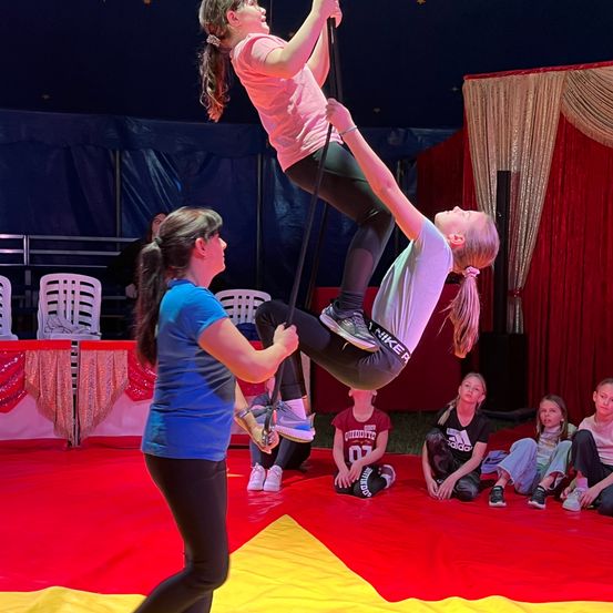 Zwei Mädchen führen einen Trapezakt in einem Zirkus auf. Ein Mädchen wird von einem anderen gehoben, während eine Menge am Boden zuschaut.